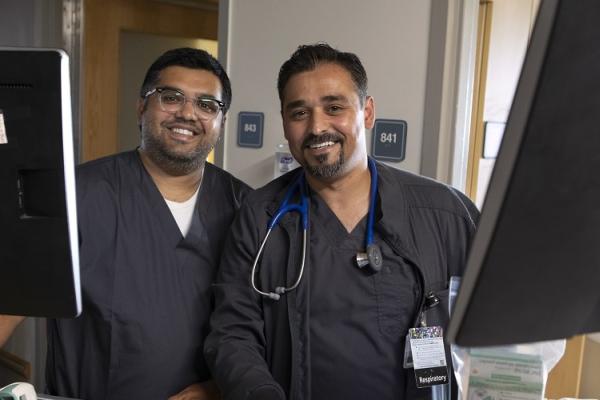 Imran Dawood, left, who is wearing glasses, and his brother, Yasin Dawood, who has a beard, stand side by side behind two medical carts with computer monitors on top. Both men are smiling and are dressed in hospital scrubs. Yasin has a stethoscope around his neck. 