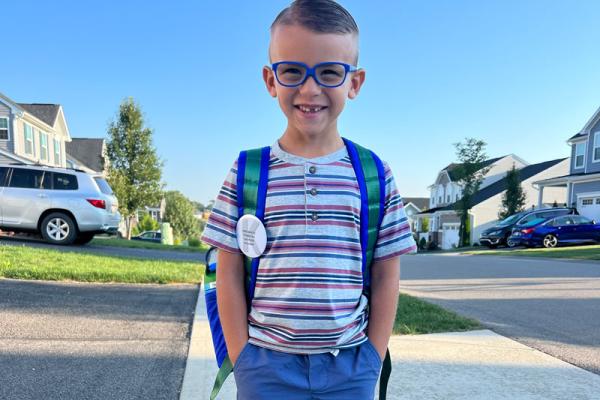 Young boy, with hands in his pockets, smiles revealing a missing tooth. He wears glasses and a backpack.