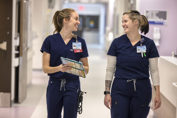Two female registered nurses smile at each other as they walk side by side down a hospital hallway. They are wearing navy blue uniforms.