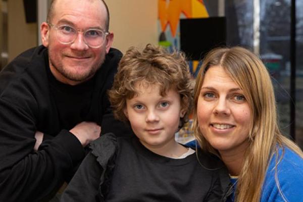 A man wearing glasses leans over the back of a chair while looking at woman kiss a little girl’s cheek. A window and monitor are visible in the background.