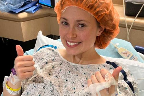 Emily is lying in a hospital bed wearing a surgical gown and hair cover, giving two thumbs up. Hospital equipment is visible in the background.