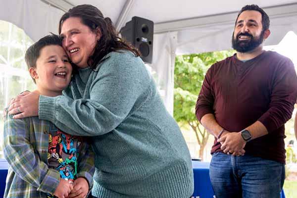 A woman leans down to embrace a young child who is standing in front of her, while an adult man stands a few feet away with his hands clasped. They are gathered inside a tented outdoor area with tables and greenery visible in the background.