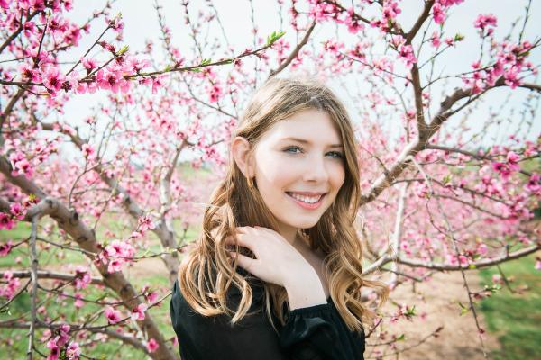 A young woman smiling, is standing in front of cherry blossom trees.