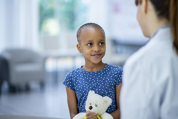A female doctor and cancer patient are indoors in a hospital room. The girl is smiling while holding a teddy bear. The doctor is telling the girl about her checkup.