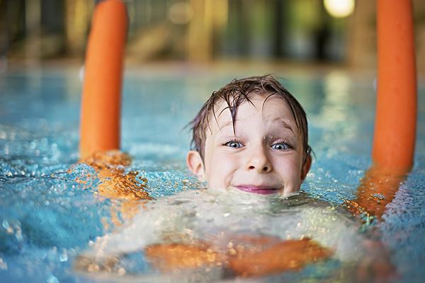 Happy kid learning to swim in indoors swimming pool. The boy is aged 5  and is using orange pool noodle. He is smiling into the camera.