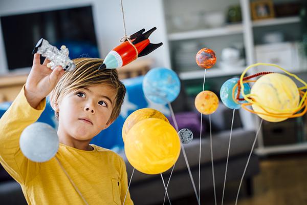 Little boy playing with his homemade planetarium as he holds an astronaut. A rocket hangs above. Arms raised as he plays.