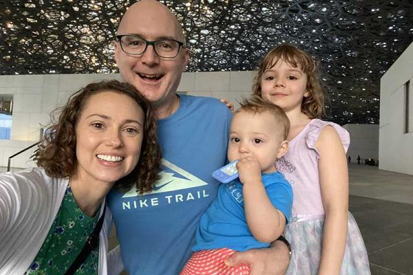A family of four takes a selfie inside a spacious modern building with a decorative, lattice-patterned ceiling overhead. One adult holds a small child while another child stands beside them.