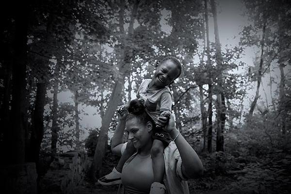 Mother smiles as son sits on shoulders and laughs while holding mother’s hands in a wooded setting.