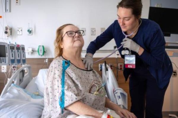 A nurse examines a hospitalized patient using a stethoscope while the patient sits upright in a hospital bed.