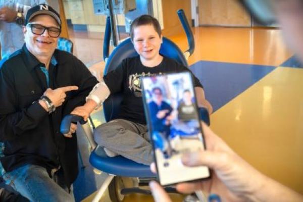 A man and a smiling young boy sit together in a hospital treatment area, with the man pointing toward the boy as another person photographs them with a smartphone.