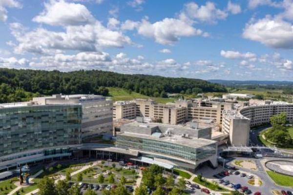 Aerial view of the Penn State Health Milton S. Hershey Medical Center campus with multiple modern buildings, parking areas, landscaped grounds, and a helipad, surrounded by wooded hills and farmland under a partly cloudy blue sky.