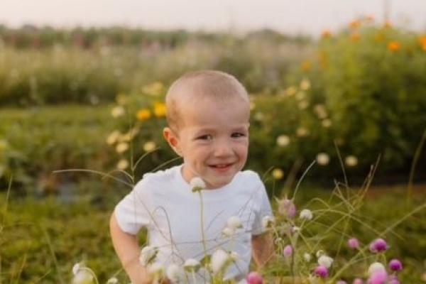 A young child wearing a white T-shirt sits among blooming wildflowers in an outdoor garden or flower field.