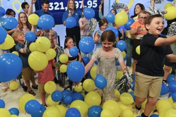 Children and event hosts celebrate during a Children’s Miracle Network fundraising reveal, tossing blue and yellow balloons into the air in front of a large display showing the total raised. The room is filled with smiling children, balloons, and pom-poms as participants cheer during the announcement.
