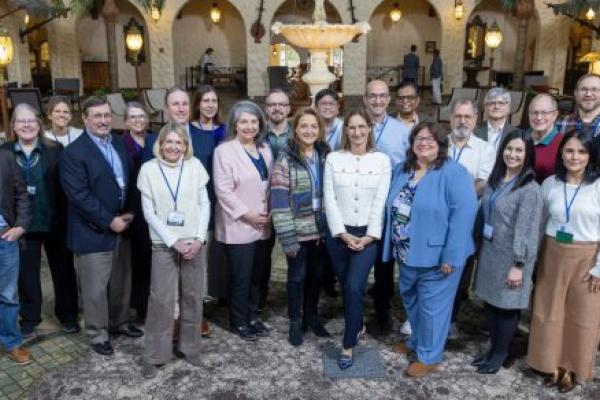 A group of approximately 23 people in business casual attire pose for a group photo in a hotel lobby. A fountain is in the near background.