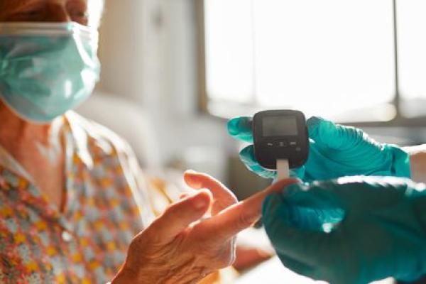 A masked woman holds her finger to a glucose meter that is held by another individual wearing gloves.