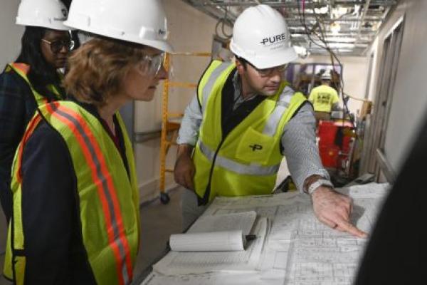 A man in a hard hat points to blueprints laid out on a table while two women in hard hats look on. They’re standing in a hallway that’s under construction.