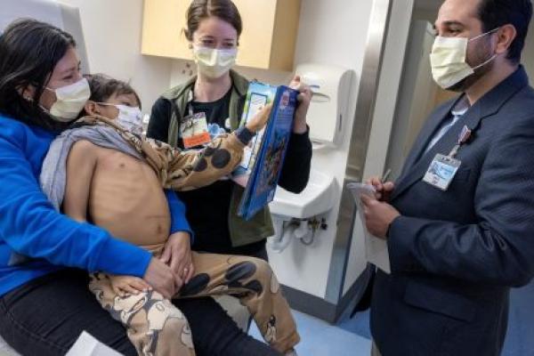 Four people in a hospital room talk behind surgical masks. One is a child, who sits on a woman's lap. The child's shirt has been pull aside. He gestures at a man who stands taking notes on a pad.