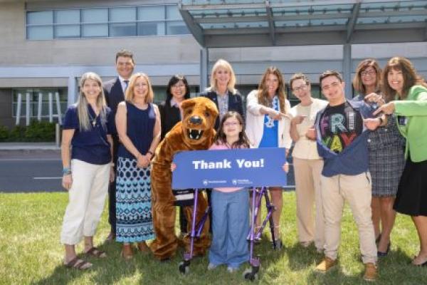 A group of people and a costumed Nittany Lion pose for a photo in front of Penn State Health Golisano Children’s Hospital, many of them smiling and pointing, “goofy pose” style. They hold a large sign that reads “Thank You!”