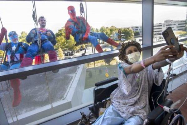 A woman in a hospital gown, cap and mask takes a selfie as three people in superhero costumes pose behind her, on the other side of a window.