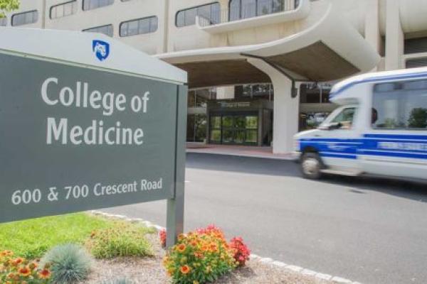 A shuttle bus drives toward the Penn State College of Medicine entrance. A sign says “College of Medicine, 600 & 700 Crescent Road.” Flowers are below the sign. The college building features rows of long windows.
