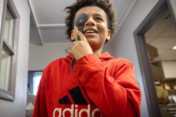 A boy holds an occluder over his right eye and smiles as he looks at a vision chart. He is wearing a sweatshirt with an Adidas logo on it.