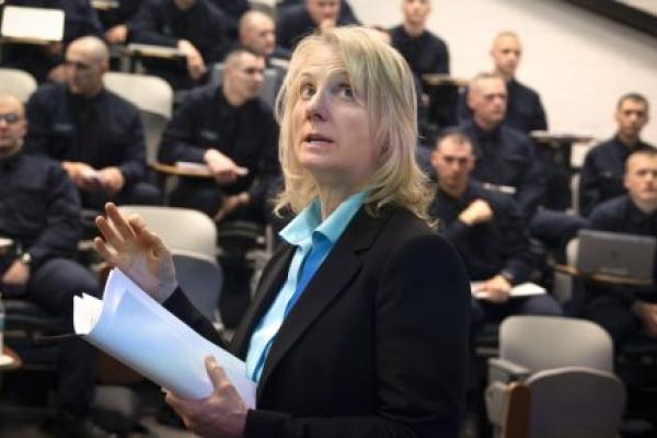 Terry Brosche of Penn State Health stands in front of a group of Pennsylvania State Police cadets who are sitting in an auditorium. She is gesturing, looking up and holding papers. She is wearing a suit jacket and blouse.