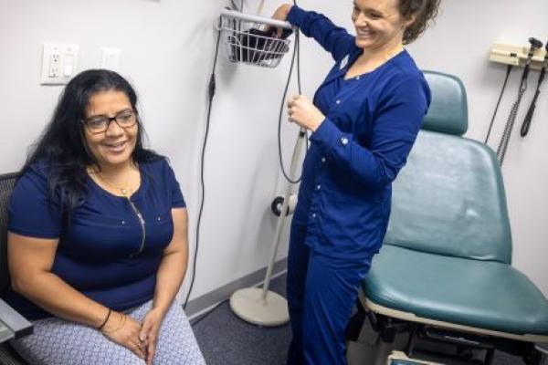 A woman patient smiles as she sits in a chair in a doctor’s exam room, while a nurse in scrubs puts a blood pressure cuff in a basket on the wall.