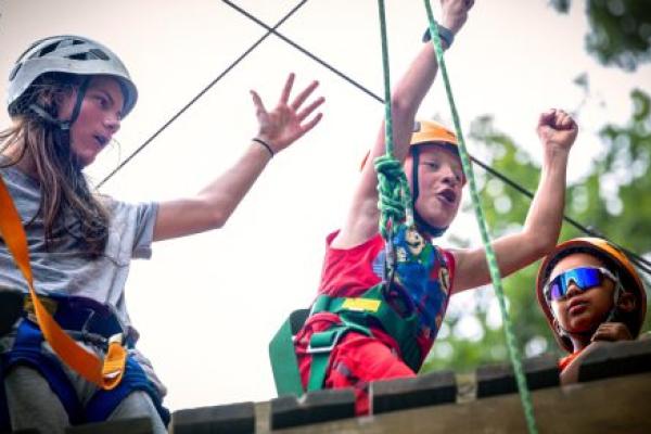 Three people stand atop a wooden platform, each wearing helmets and safety equipment related to ziplining. The child in the middle has his hands raised in the air.