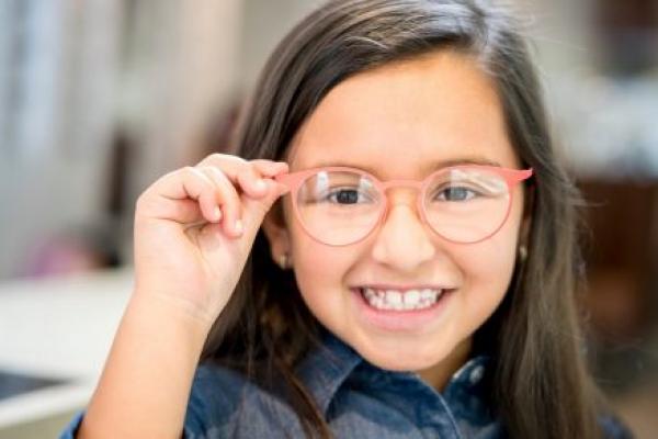 A young girl tries on glasses, smiling toward the camera.