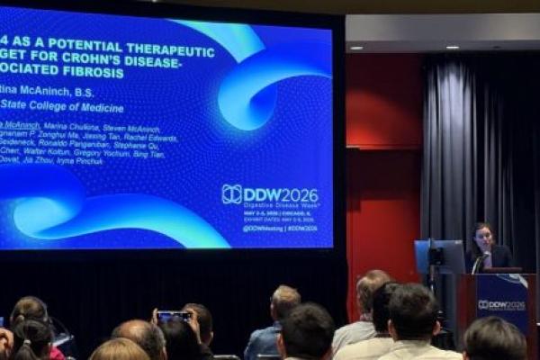 A person stands at a podium next to a large screen displaying a research presentation in front of a large conference room.
