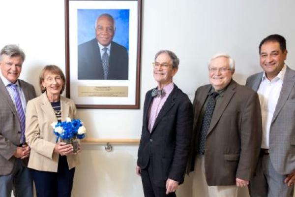 Richard Legro, MD; Cecile Mortel; Joel Sorosky, MD; Raymond Hohl, MD, PhD; and Mark Shahin, MD, stand together smiling beside a portrait of longtime College of Medicine faculty member and humanitarian Rodrigue Mortel, MD, at Penn State Cancer Institute.