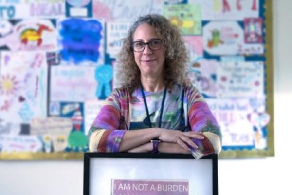 Amy Keisling, Gender Health Clinic coordinator, smiles as she leans on a framed picture that says “I am not a burden.” She has curly hair and is wearing glasses and a lanyard. Behind her is a bulletin board out of focus.