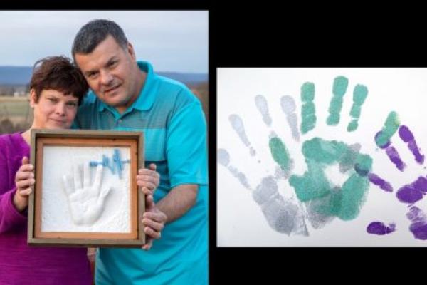 Wendy Gilbert, who has short hair and wears a bright top and jean, and Ronald Gilbert, who wears a polo shirt and jeans, hold a framed shadow box containing a resin handprint and 3D heartbeat. They are standing outside with a field in the background. Dylan’s handprints are beside them.