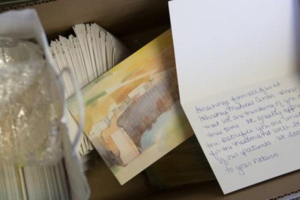 Stacks of cards sit in a cardboard box. The front of the card shows a watercolor painting of Hershey Medical Center. One card is propped open to show handwriting inside.