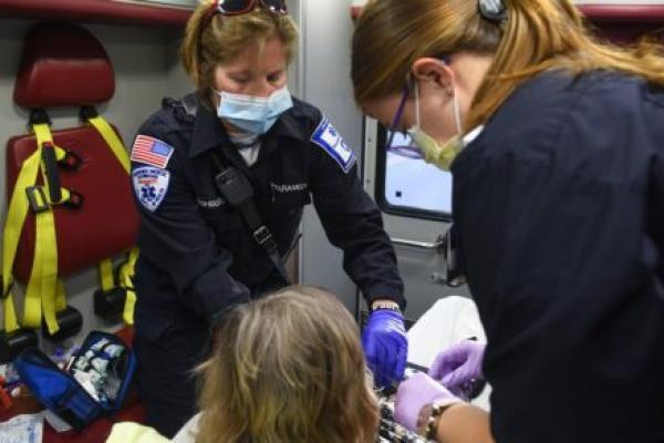 Paramedic Sarah Fishburne, who has short hair with glasses on top of her head and wears a face mask, and EMT Madigan Anderson, who has her hair in a ponytail and wears glasses and a face mask, sit in an ambulance with a patient on a stretcher, who is seen from behind. Medical equipment surrounds the patient.
