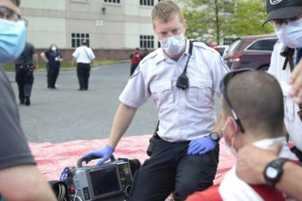 Four men are circled around a piece of equipment in a parking lot. They wear surgical masks. Two wear uniforms. Behind them other men and women in uniform stand in the parking lot.