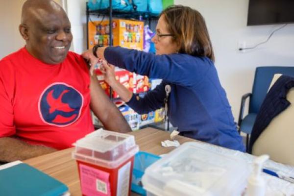 A man in a t-shirt smiles as a nurse gives him a flu shot in his left arm.