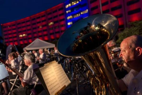 A man plays a tuba in the foreground while other musicians are in the background, in front of a crescent-shaped building lit up in red and blue lights.