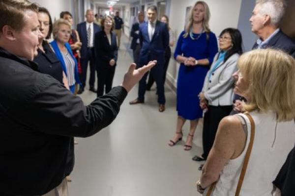 A group of about 15 people wearing professional attire stand in the hallway of a newly refurbished hospital unit. A man in the foreground has his arm extended outward as he speaks to the group.