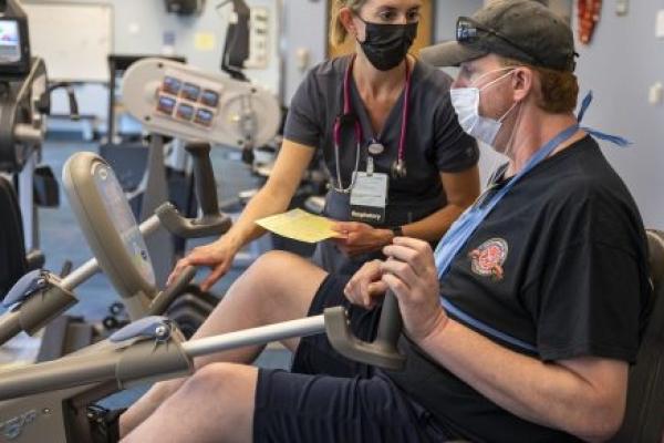 A man in a ball cap and shorts works on a piece of equipment in a gym. A woman in a surgical mask stands next to him.