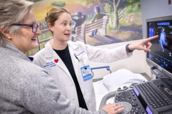 A doctor wearing a physician’s coat points to a computer screen with an ultrasound image on it. Another clinical staff member looks on. A wall in the near background has a mural of two people sitting on a bench looking out over a body of water.