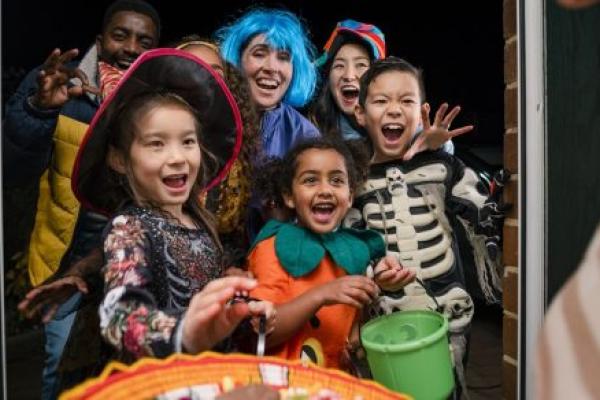 A group of children in Halloween costumes hold trick-or-treat buckets as they stand on a front porch doorway. A portion of a candy dish held by an adult is in the foreground.