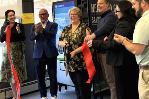 Six people smile and clap immediately after cutting a ribbon, as they stand in front of a vending machine. A window is also in the background.
