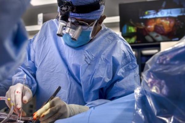 A surgeon wearing a blue surgical gown, mask, and magnifying loupes performs a heart transplant surgery in an operating room, using instruments while monitors display the procedure in the background.