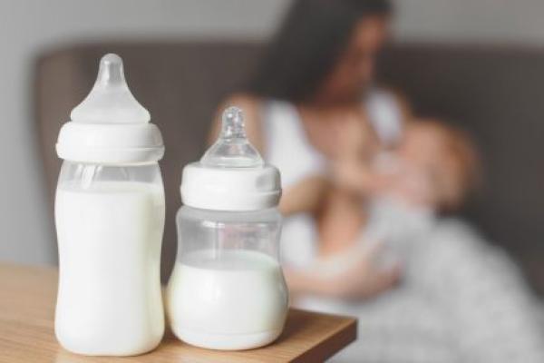 Two bottles of breast milk sit on a counter. Faded into the background, a mother breastfeeds her baby.