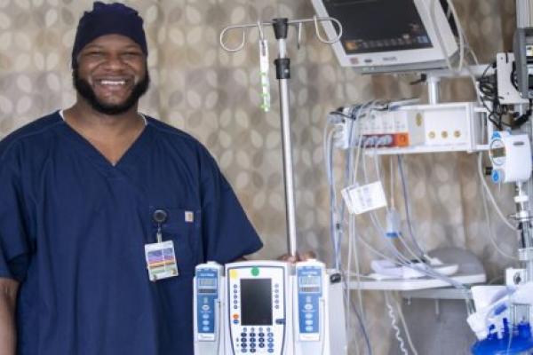 Nurse Viniquinn Terry, who has a beard and wears scrubs, smiles as he stands with medical equipment used in the cardiovascular intensive care unit.