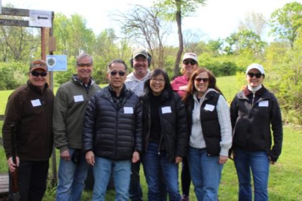 Eight people wearing casual outdoor attire pose for a photo. The man on the far left is posing with a rifle. Grass, bushes and trees are in the background.