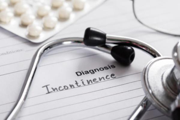 A piece of paper with the words "Diagnosis Incontinence" sits on a desk. On top of it are a stethoscope, pack of pills and reading glasses.