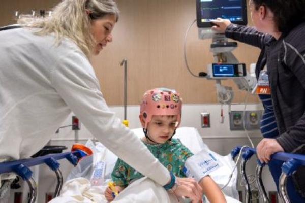 A young child wearing a monitoring cap sits on a hospital bed while two medical staff assist with care and adjust medical equipment.