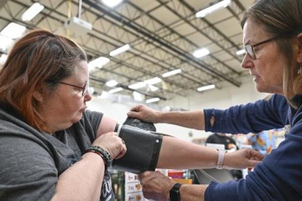 A woman holds a blood pressure cuff on her arm as a nurse on the right adjusts it. They are sitting at a table in a large warehouse room with a high ceiling and bright fluorescent lights.